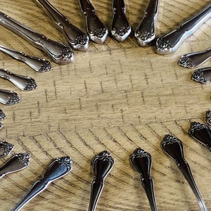 May include: An assortment of vintage silverware arranged on a wooden surface. The silverware includes forks, spoons, and knives with ornate floral detailing. The arrangement creates a circular pattern, showcasing the elegant design.