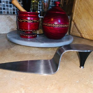May include: A stainless steel cake server with a unique design, featuring a curved handle and a black heel-shaped base. The server is placed on a light brown surface, with a red wooden mortar and jar in the background.