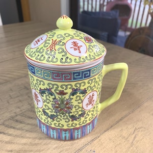May include: Yellow ceramic mug with a lid, featuring traditional Chinese designs. The mug has a handle and is decorated with floral patterns, Chinese characters, and a blue and red border. The lid is topped with a small yellow knob.