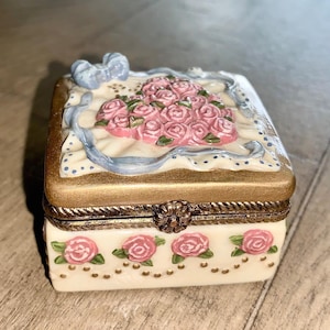 May include: A small, rectangular trinket box with a hinged lid. The box is cream-colored with gold trim and features a bouquet of pink roses on top. The sides are decorated with more pink roses and gold accents. A blue bow adorns the top.