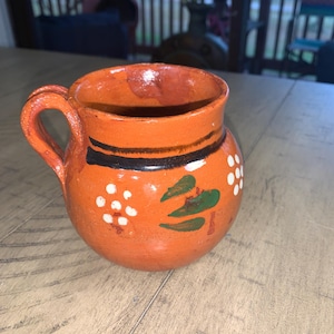 May include: A hand-painted, orange ceramic mug with a handle. The mug features black and white bands around the rim and floral designs in white and green. The mug is likely handmade and suitable for hot beverages.