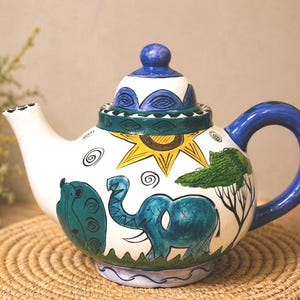 May include: A hand-painted ceramic teapot with a blue handle and lid. The white body features a sun, elephant, and tree design in blue, green, and yellow. The teapot sits on a woven mat.