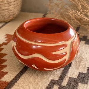 May include: A round, terracotta-colored ceramic bowl featuring a smooth, polished finish and a wide, open top. The exterior is decorated with carved, cream-colored abstract wave patterns that wrap around the circumference of the vessel.