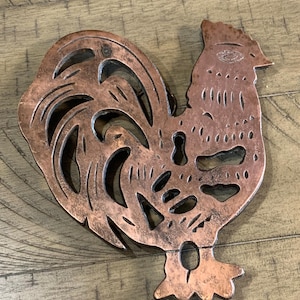 May include: A copper-colored metal trivet in the shape of a rooster. The trivet has a detailed design with cut-out sections in the tail and body. The rooster is positioned on a wooden surface.