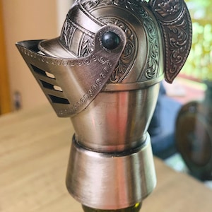 May include: A decorative bottle stopper shaped like a knight's helmet. The silver-colored metal helmet features intricate embossed details and a hinged visor. The stopper is designed to fit on a wine bottle.