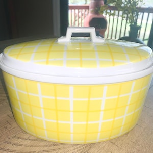 May include: A yellow and white ceramic casserole dish with a lid. The dish has a grid pattern and a white handle on the lid. The dish is sitting on a wooden table.