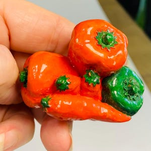 May include: A handcrafted, miniature sculpture of colorful peppers. The sculpture features a vibrant orange bell pepper, a red chili pepper, and a green bell pepper, all with detailed green stems.