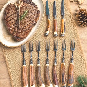 May include: A cooked T-bone steak on a white plate with a sprig of rosemary. The image also shows a set of steak knives and forks with antler handles, arranged on a burlap placemat. A pine cone and greenery are in the background.