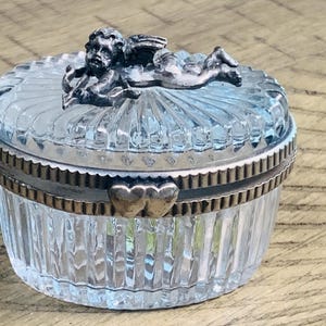 May include: A round, clear glass trinket box with vertical ribbing on the sides and lid. The lid features a metallic silver-toned cherub figurine on top. A metallic silver-toned band with a heart-shaped clasp secures the lid to the base.