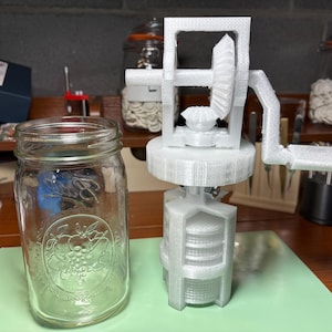 May include: A clear glass jar with a decorative emblem and a white 3D-printed mechanical device with a crank handle. The device has gears and a cylindrical base. The objects are on a green surface.