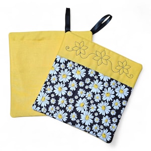 May include: Two square potholders with black hanging loops. One is solid yellow with black embroidered flower outlines. The other features a black fabric panel with a white daisy pattern and a yellow top border with black embroidered flowers.