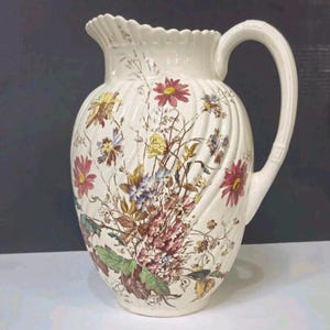 May include: A cream-colored ceramic pitcher with a scalloped rim and a ribbed body. The surface is decorated with a colorful botanical print featuring various wildflowers in shades of red, yellow, blue, and green, complete with a curved handle.