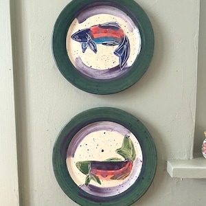May include: Two round ceramic plates featuring hand-painted fish designs. Each plate has a wide dark green rim and a cream-colored center with purple brushstrokes and black speckles. The top plate shows a blue and orange fish, while the bottom plate displays a green and orange fish.