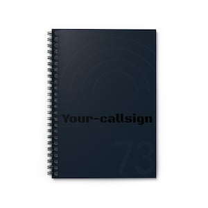 Puede incluir: Un cuaderno de espiral azul oscuro con el texto "Your-callsign" en negro. El número "73" está impreso en un tono de azul más claro. La portada presenta un diseño circular sutil.