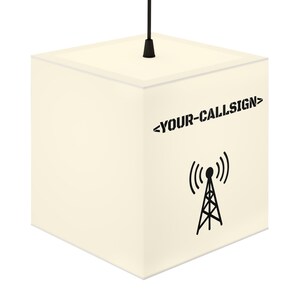 Puede incluir: Lámpara colgante en forma de cubo de color crema. La lámpara presenta el texto "<YOUR-CALLSIGN>" y un gráfico negro de una torre de radio con ondas de señal. La lámpara cuelga de un cable negro.