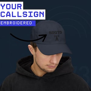 May include: Navy blue baseball cap with embroidered text "SIG73" and a radio tower graphic. The hat also features the text "YOUR CALLSIGN EMBROIDERED" in white. A classic, adjustable hat.