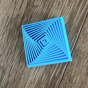 Puede incluir: Un objeto cuadrado azul brillante con un diseño en espiral concéntrico. El objeto está hecho de un material sólido y descansa sobre una superficie de madera. El diseño crea un efecto 3D. Dimensiones: aproximadamente 5 cm x 5 cm.