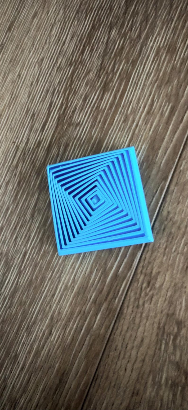 May include: A bright blue, square-shaped object with a concentric, spiral design. The object is made of a solid material and sits on a wooden surface. The design creates a 3D effect.