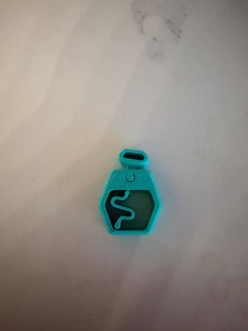 Puede incluir: Un colgante hexagonal turquesa con un dise&ntilde;o de serpiente en el interior. El colgante tiene un peque&ntilde;o lazo en la parte superior y un detalle en forma de bot&oacute;n. El dise&ntilde;o de la serpiente es una l&iacute;nea simple y curvada. El colgante es probablemente de pl&aacute;stico o resina.