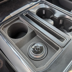 Peut inclure: Gros plan sur la console centrale d'une voiture, avec un sélecteur de vitesse rotatif affichant les positions P, R, N et D. La console comprend des porte-gobelets et un compartiment de rangement, en noir et gris.