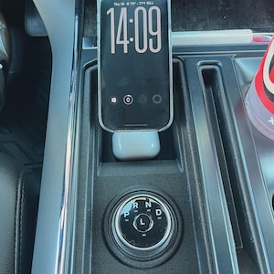 Peut inclure: Un smartphone affichant 14h09 est posé dans la console d'une voiture, au-dessus d'écouteurs sans fil blancs. La console comprend également un sélecteur de vitesse avec les positions P, R, N, D et L.