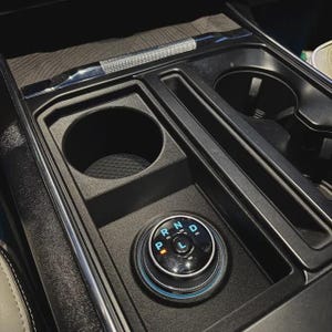 May include: A black automotive center console featuring a circular gear shift dial with illuminated blue letters P, R, N, D, and L. The console includes a textured cup holder, a narrow storage slot, and a metallic trim accent along the top edge.