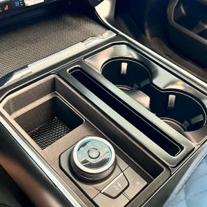 May include: A black automotive center console featuring a circular gear shift dial with illuminated P, R, N, D, and M letters. The console includes two circular cup holders, a rectangular storage compartment with a textured base, and a small button.