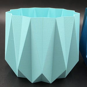 Puede incluir: Una maceta geométrica azul claro con un diseño facetado. La maceta tiene forma hexagonal y está hecha de un material texturizado. Los lados están angulados, creando un aspecto moderno. Ideal para plantas pequeñas o suculentas.
