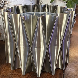 Puede incluir: Un recipiente decorativo plateado con un diseño geométrico en forma de diamante. El recipiente tiene un inserto de vidrio transparente y está hecho de metal plegado. El recipiente está diseñado para contener objetos y es adecuado para la decoración del hogar.