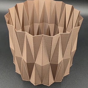 Puede incluir: Una maceta geométrica marrón con un diseño facetado. La maceta tiene una amplia abertura y una superficie texturizada, con una serie de formas triangulares que crean su forma. La maceta está hecha de un material sólido.