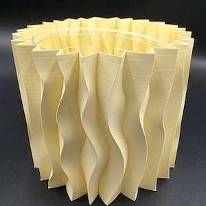 May include: A cream-colored, accordion-style lampshade with a wavy, pleated design. The shade has a cylindrical shape and a clear plastic ring at the top. The texture of the shade appears to be paper or fabric.