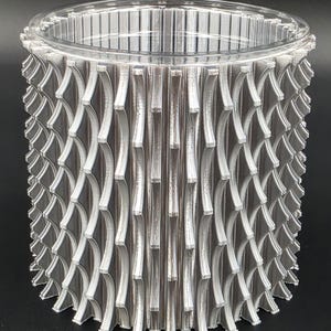 May include: A cylindrical, silver-colored object with a clear, ridged top. The body features a repeating pattern of curved, vertical elements, creating a textured surface. The design is symmetrical and modern, with a focus on geometric shapes.