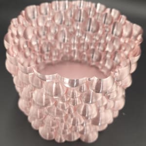 Puede incluir: Un recipiente decorativo de color rosa claro con un patrón texturizado en forma de burbujas. El recipiente tiene un borde festoneado y una forma cilíndrica, adecuado para guardar objetos pequeños o como pieza decorativa. El recipiente es de material rosa claro.