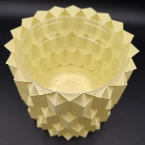 May include: A light yellow geometric planter with a clear plastic liner. The planter has a repeating pattern of diamond shapes, creating a textured surface. The top edge has a series of triangular points. Ideal for small plants or succulents.