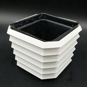 Puede incluir: Una maceta cuadrada blanca y negra con un diseño único en capas. La maceta tiene un interior negro y un exterior blanco con una serie de secciones angulares apiladas. La forma general es geométrica y moderna.