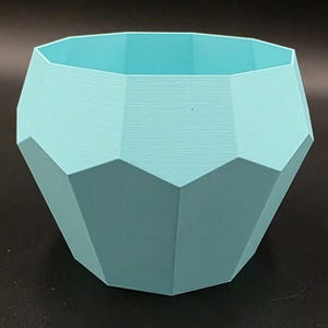 Puede incluir: Una maceta geométrica azul claro con un diseño facetado. La maceta tiene una abertura ancha y una superficie texturizada. Este recipiente decorativo es adecuado para plantas pequeñas y suculentas.