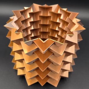 Puede incluir: Una escultura geométrica y en capas con un diseño simétrico. El objeto está compuesto por numerosos elementos triangulares y en forma de V en tonos bronce y beige, creando una forma texturizada y tridimensional. El interior revela un espacio hueco.