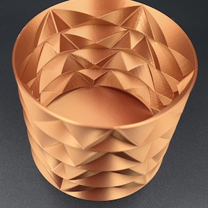 Puede incluir: Un recipiente cilíndrico de color cobre con un exterior geométrico con forma de diamante. El interior es liso y el recipiente tiene una abertura ancha. El diseño presenta formas triangulares repetidas, creando una superficie texturizada. El recipiente mide unos 10 cm de alto.