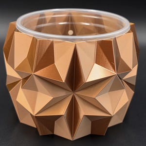 May include: A geometric, bronze-colored planter with a clear plastic liner. The planter features a starburst design on the front and is made of a series of triangular shapes. The planter is designed for indoor or outdoor use.