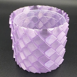 Puede incluir: Un recipiente de color lavanda con un patrón tejido. Tiene un inserto cilíndrico transparente. El exterior presenta un patrón de diamantes repetido, creando una superficie texturizada. El diseño es moderno y geométrico.