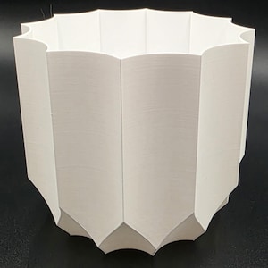 Puede incluir: Una maceta geométrica blanca con un borde festoneado. La maceta tiene una serie de paneles verticales en ángulo que crean una superficie texturizada. El diseño es moderno y minimalista, adecuado para uso en interiores o exteriores.