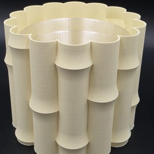 May include: A cream-colored, cylindrical vase with a bamboo-inspired design. The vase features a scalloped top edge and a series of vertical, rounded segments that mimic bamboo stalks. The interior is smooth and the exterior has a textured finish.
