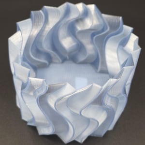Puede incluir: Un cuenco decorativo azul claro impreso en 3D con un diseño ondulado único. El cuenco tiene forma cilíndrica con un exterior texturizado y en capas. El interior es liso y el cuenco está hecho de un material translúcido.