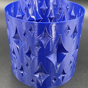 May include: A vibrant blue cylindrical container with a repeating diamond pattern. The container is made of a solid material and has a smooth, reflective surface. The diamond shapes are raised, creating a textured effect. The container is empty and sits on a dark surface.