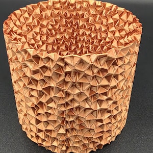 Puede incluir: Un recipiente cilíndrico de color cobre con una superficie texturizada compuesta por numerosas formas triangulares. El diseño geométrico crea un patrón único y llamativo. El recipiente es probablemente un artículo decorativo o un pequeño recipiente de almacenamiento.