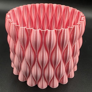 Puede incluir: Una maceta rosa, impresa en 3D, con un diseño tejido y texturizado. La maceta cilíndrica presenta un borde superior festoneado y patrones verticales en forma de lágrima. El color es un rosa suave y apagado.