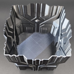 May include: A 3D-printed, geometric container with a square shape and a detailed, textured design. The container is primarily dark gray with lighter gray accents, and features a symmetrical pattern on the sides and interior.