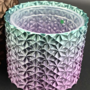 May include: A cylindrical planter with a geometric pattern in shades of green, purple, and silver. The planter has a clear plastic insert and a textured exterior. The design features a repeating starburst pattern. The planter is suitable for indoor or outdoor use.