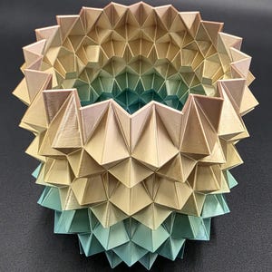 May include: A 3D-printed geometric vase with a layered design. The vase features a gradient of colors, transitioning from gold to teal. The intricate structure is composed of repeating triangular shapes, creating a unique decorative piece.
