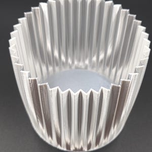 Puede incluir: Un recipiente transparente y acanalado con un borde en zigzag. El recipiente está hecho de un material translúcido y tiene un interior gris claro. Las crestas crean un patrón vertical alrededor de los lados del recipiente.